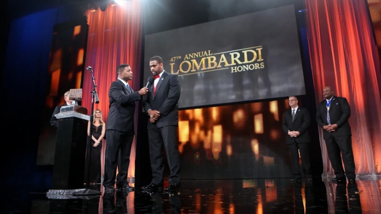 Two men speaking on stage at the 4th annual lombardi honors event, with attendees watching in the background.
