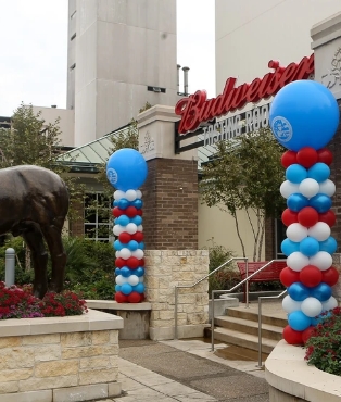 Decorative blue and red balloons flank an entrance with a budweiser sign above, near a bronze statue of a bull.
